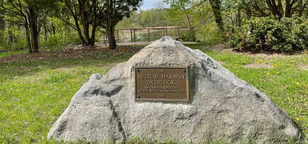 A large rock with a bronze plaque reading "The Reuel D. Harmon Overlook" with a deck and pond in the background.